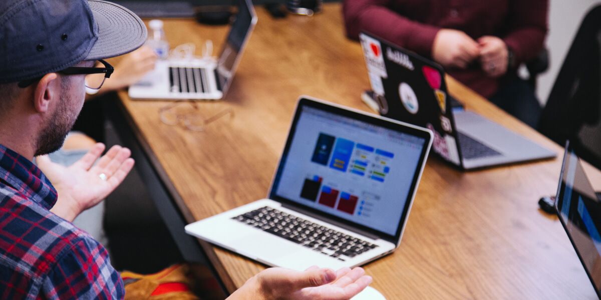 A photo of group of people around a table with laptops.  The person in the foreground has his hands upraised in a shrugging gesture, as if he is asking a question.